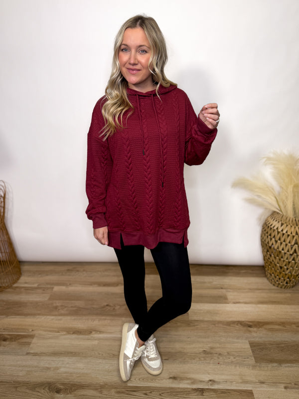 Woman wearing a burgundy cable knit hoodie in a room with wooden flooring and decorative items.