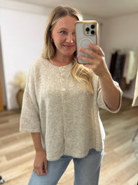 A woman taking a mirror selfie in a boutique or home setting, wearing a light beige knit cardigan sweater with subtle multicolor flecks and light blue jeans.