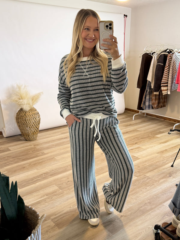 A woman takes a mirror selfie in a boutique while wearing a matching two-piece loungewear set with horizontal green and white stripes, featuring a long-sleeve top and wide-leg pants with a white drawstring.