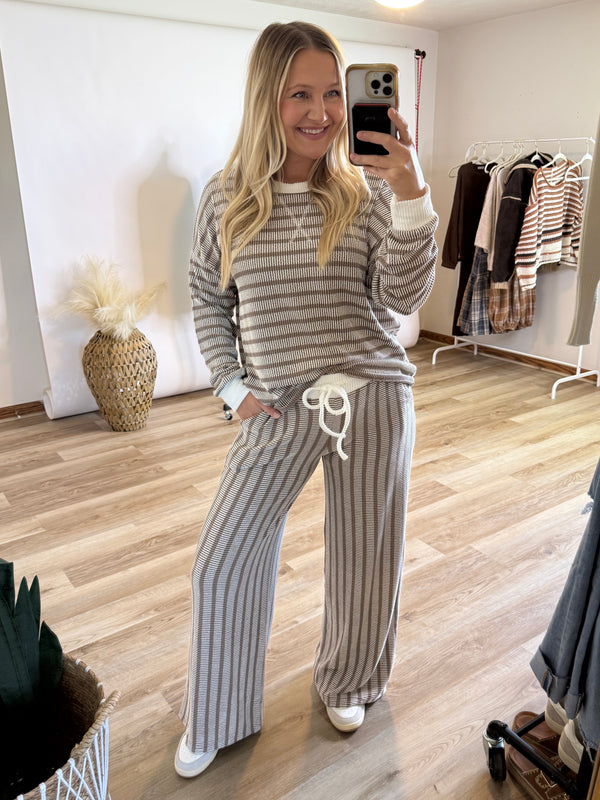 A woman takes a mirror selfie in a boutique showing a matching two-piece loungewear set consisting of a long-sleeve striped top and wide-leg pants. The set features a beige vertical stripe pattern with a white drawstring waist.