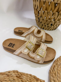 Beige sandals with gold buckles on a white surface with woven baskets.