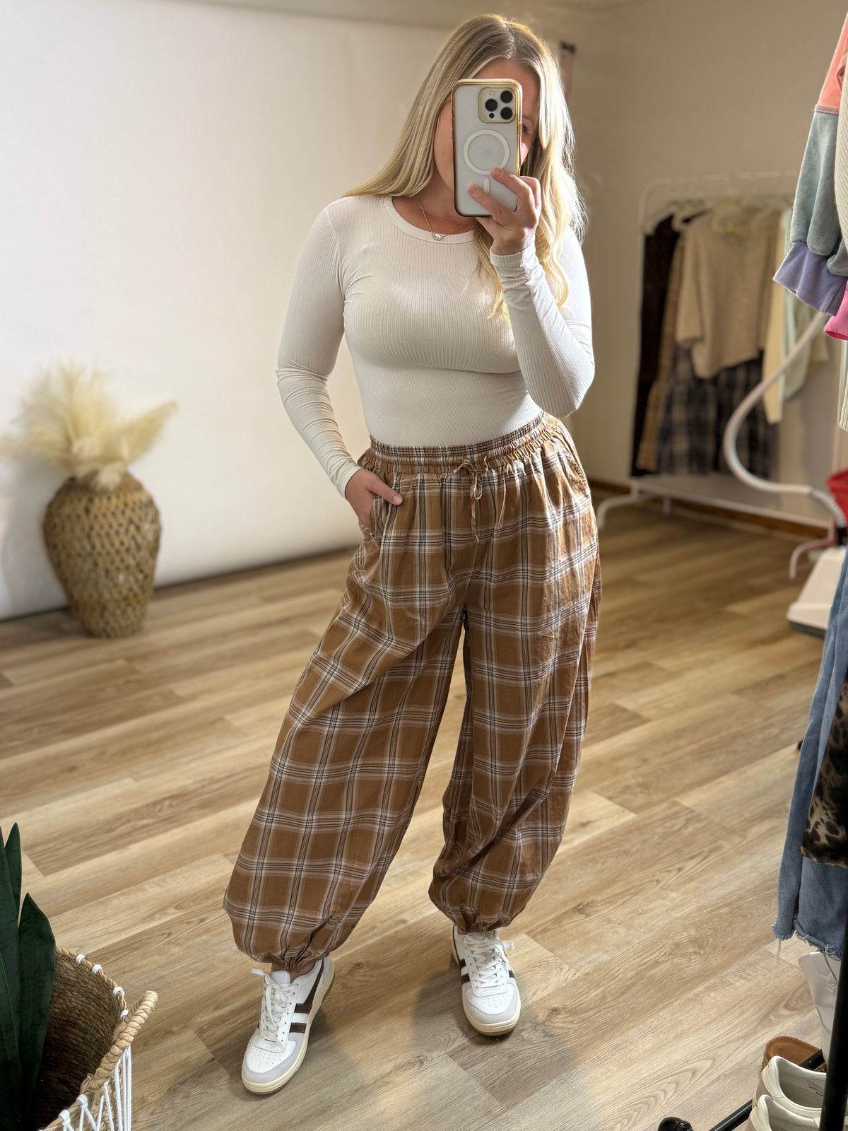 A woman takes a mirror selfie in a room, wearing a fitted white long-sleeve top and brown plaid wide-leg pants with white sneakers.