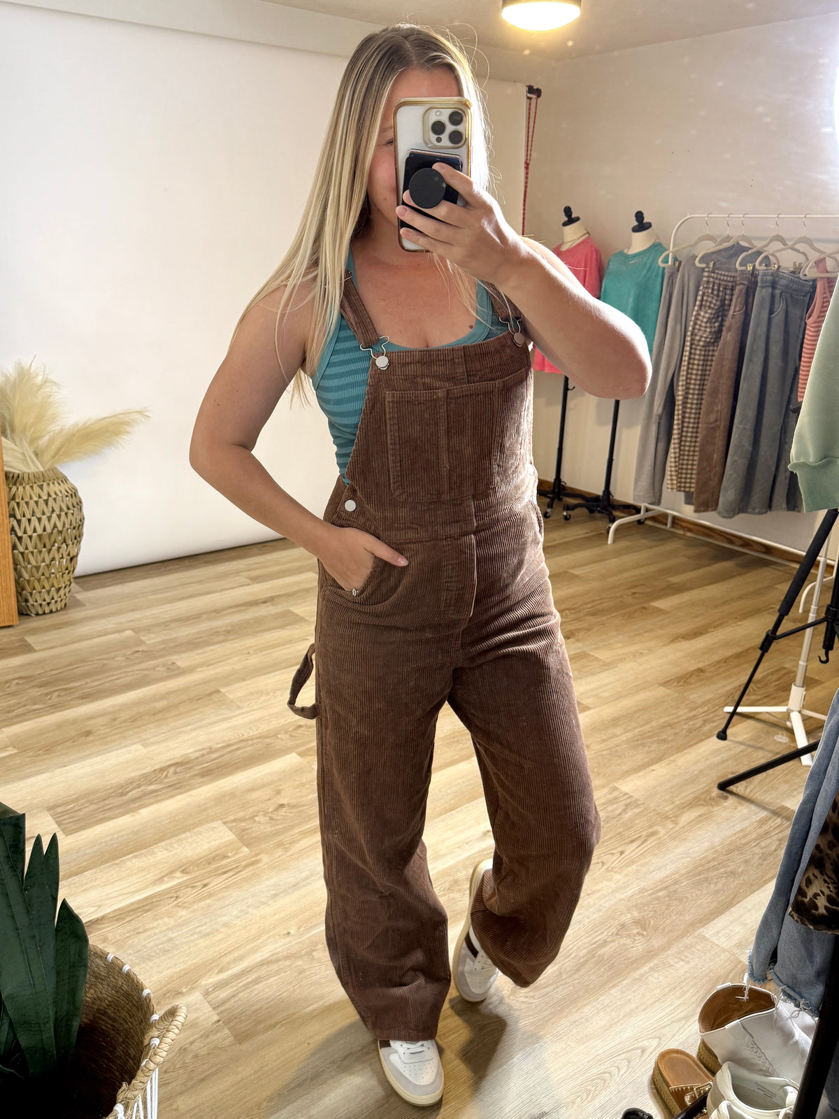 A woman takes a mirror selfie in a boutique wearing brown corduroy overall sweatpants over a teal striped tank top and white sneakers.