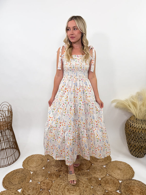 Wildflower Tiered Smocked Maxi Dress with Self Tying Straps – White with watercolor wildflower prints, smocked stretchy top, tiered flowy skirt, self-tie adjustable straps, side pockets, and fully lined. Perfect for spring and summer occasions like Easter, garden picnics, and baby showers. Flattering and boho-chic.